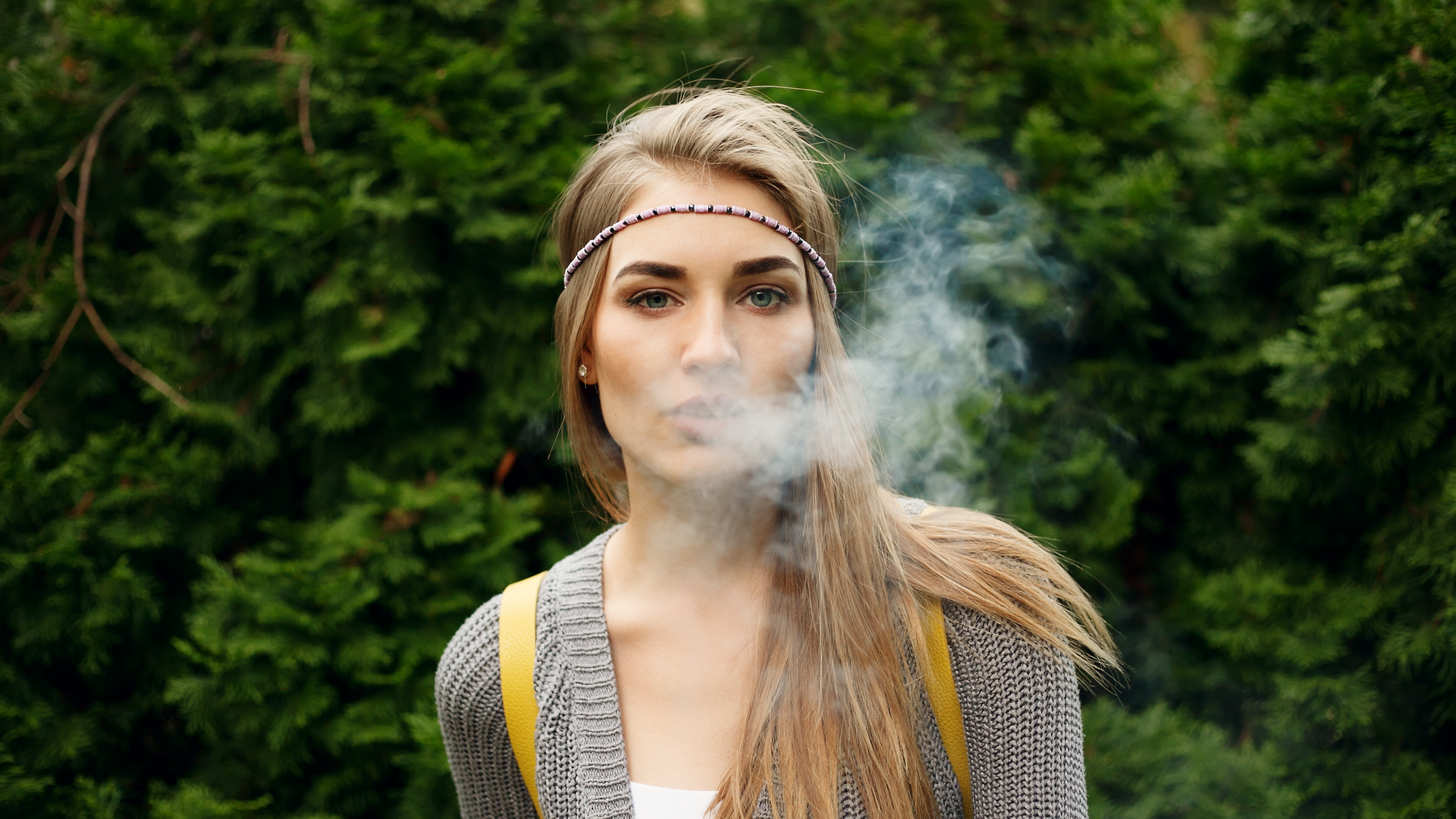 A blonde woman with a headband exhaling smoke outdoors with foliage in the background
