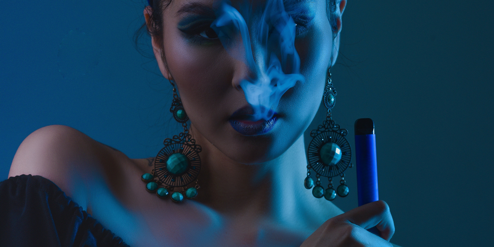 Woman holding an all in one disposable vape while exhaling smoke from her mouth with a blue hue lighting